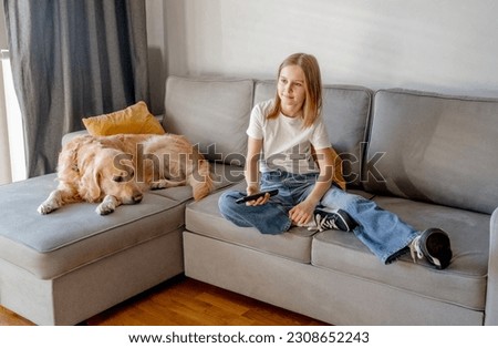 Preteen girl with golden retriever dog holding tv remote control and sitting on sofa at home. Beautiful child kid with labrador doggy pet watching cartoons at television