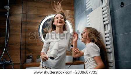 Funny young woman and 10 years old girl with long hair in pajamas drying her voluminous hair by hair dryer while dancing laughing and looking at each other in modern bathroom in home Royalty-Free Stock Photo #2308687735