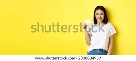 Young asian woman in casual white t-shirt showing plastic credit card and smiling at camera, yellow background.