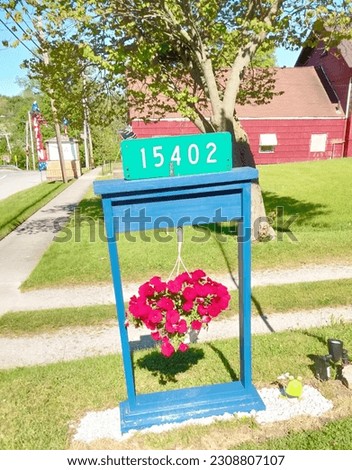 A hanging basket with red flowers is part of a decorative address marker. There is a red barn and a tree in the background. 