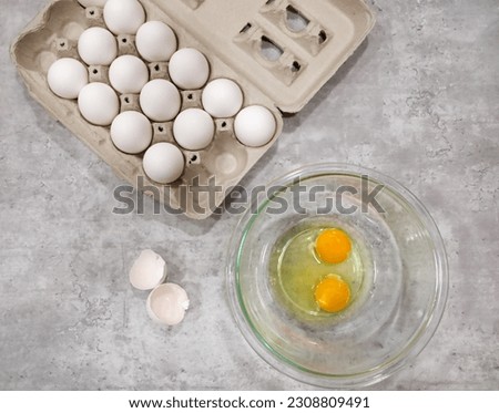 Top view of two cracked eggs in a glass bowl, eggshells and an egg carton on the side.