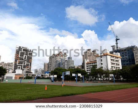 Photo of a hostel building with several apartment buildings seen from the sidelines of a soccer field with a running track on a sunny day.