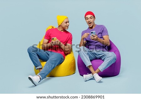 Full body cheerful happy happy young couple two friend men wear casual clothes together sit in bag chair hold in hand use mobile cell phone isolated on pastel plain light blue cyan background studio Royalty-Free Stock Photo #2308869901