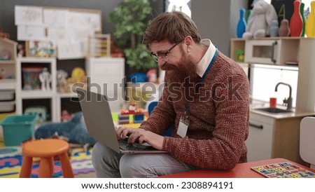 Young redhead man preschool teacher using laptop sitting on chair at kindergarten