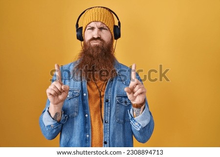 Caucasian man with long beard listening to music using headphones pointing up looking sad and upset, indicating direction with fingers, unhappy and depressed. 
