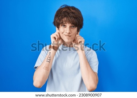 Hispanic young man standing over blue background smiling pulling ears with fingers, funny gesture. audition problem 