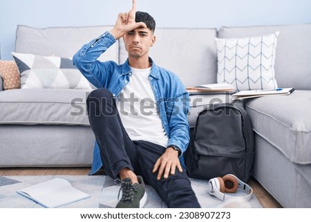 Young hispanic man sitting on the floor studying for university making fun of people with fingers on forehead doing loser gesture mocking and insulting. 