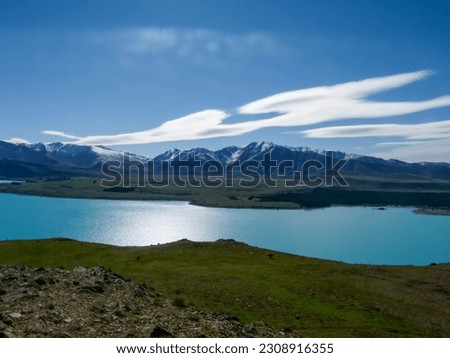 The photo shows the blue Lake Tekapo and the snow-capped mountain peaks of the Southern Alps on a sunny day with blue sky on the South Island of New Zealand.