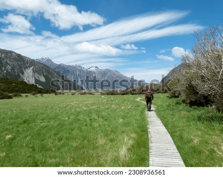 The picture shows a hiker on a boardwalk in Aoraki Mount Cook National Park on the South Island of New Zealand on a sunny day with blue sky and a few clouds.