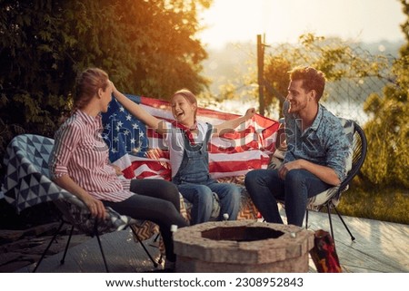  A joyous family celebrating Independence Day outdoors. The image features a mother, father, and daughter seated around a cozy fire pit, their faces beaming with laughter and delight.