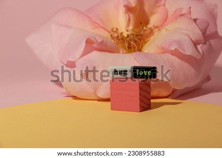 pink cube with hug and love tiles, behind the cube is a pink rose, a creative romantic design on a pink-yellow background