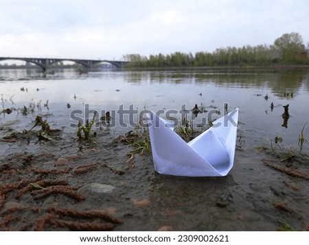 Soaked paper boat on the banks of the Yenisei River. Close-up of white paper boat on river at gloomy morning. Concept background of children dreams, hopes and wishes