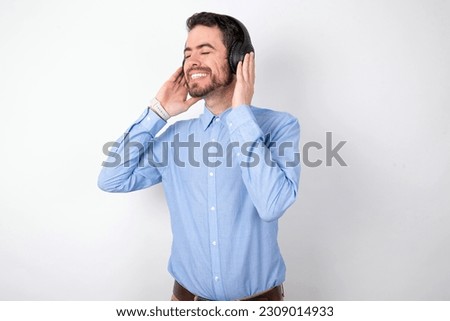 handsome businessman wearing blue shirt over white studio background with headphones on her head, listens to music, enjoying favourite song with closed eyes, holding hands on headset.