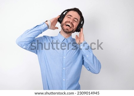 handsome businessman wearing blue shirt over white studio background smiles broadly feels very glad listens favourite music track via wireless headphones closes eyes.