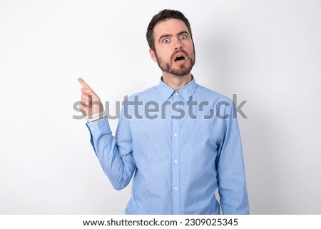 Emotive handsome businessman wearing blue shirt over white studio background keeps jaw dropped from shock demonstrates amazing promo points right on blank space demonstrates big shopping sale.