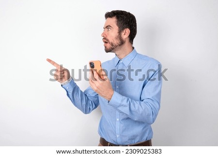 Stunned handsome businessman wearing blue shirt over white studio background points sideways right copy space, recommends product, sees astonishing thing