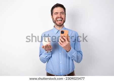 handsome businessman wearing blue shirt over white studio background holding piggy bank taking a selfie  celebrating success