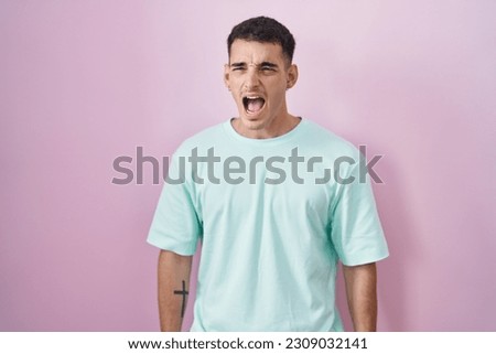Handsome hispanic man standing over pink background angry and mad screaming frustrated and furious, shouting with anger. rage and aggressive concept. 