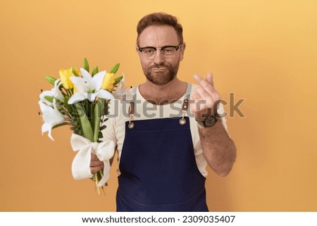 Middle age man with beard florist shop holding flowers doing money gesture with hands, asking for salary payment, millionaire business 