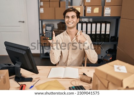 Young man working at small business ecommerce holding dataphone smiling happy and positive, thumb up doing excellent and approval sign 