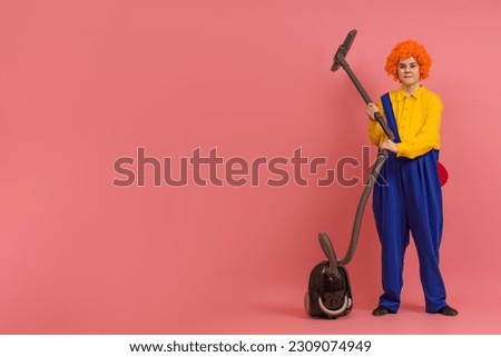 young woman in a wig and a clown costume with a vacuum cleaner on a colored background with copy space