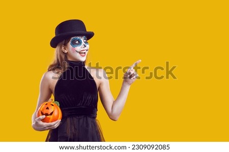 Joyful preteen girl pointing her finger at empty ad space recommending something on Halloween theme. Smiling child with halloween make-up and small pumpkin shows copy space on orange background.