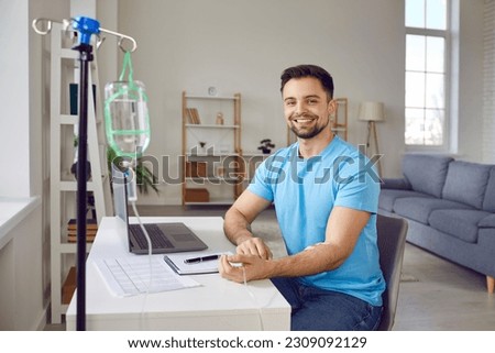 Portrait of happy, smiling, young man patient receiving intravenous vitamin infusion through sterile IV line in his arm while sitting at desk with laptop computer at home. Vitamin therapy concept Royalty-Free Stock Photo #2309092129