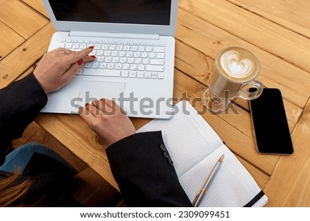 Female hands with red manicure on the background of a white laptop. The girl in the cafe works on a laptop. On the table is a cup of coffee, a diary and a phone next to a laptop