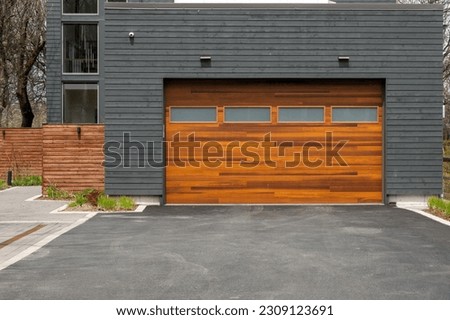 A modern brown faux wooden exterior garage door with four small horizontal glass windows. The modern door is on a luxury dark grey contemporary house with a concrete driveway.  Royalty-Free Stock Photo #2309123691