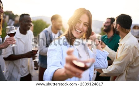 Point of view of pretty young woman toasting with glass of red wine in hand and smiling. Girl posing for photo at party with her friends outdoors at sunset. People gathered celebrating success. Royalty-Free Stock Photo #2309124817