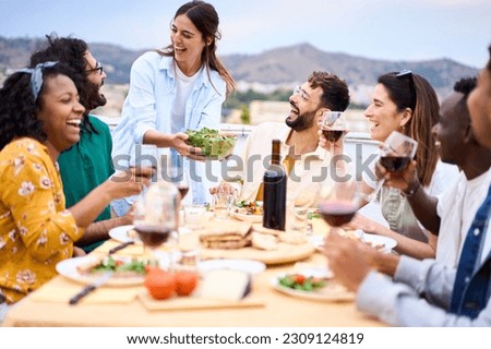Young smiling Caucasian woman putting plate with food on table at celebration with happy friends on rooftop. Group of cheerful multiracial friends gathered for barbecue party on outdoor terrace.