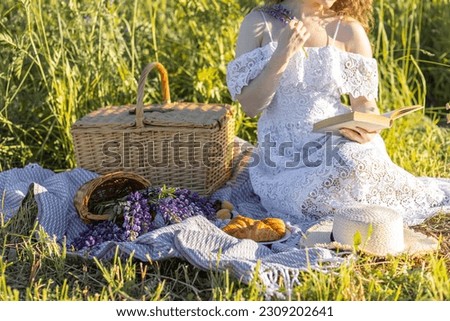 Beautiful young girl in a white dress, straw hat, picnic basket reading a book on a meadow. Summertime, golden hour, sunset. Work life balance concept
