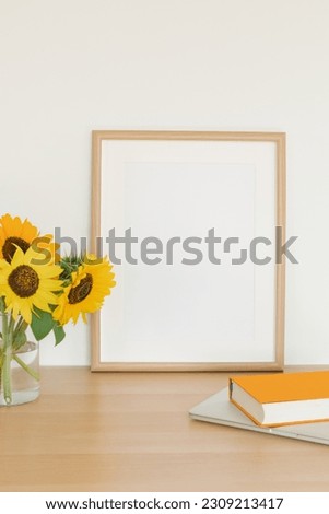 Vase with sunflowers next to a blank picture frame, a hardcover book, laptop. Office desk. Poster frame mockup concept.