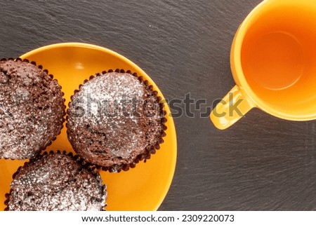 Three homemade chocolate muffins with ceramic yellow saucer and cup on slate stone, macro, top view.