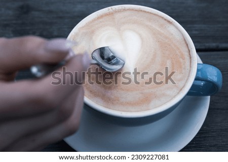 Blue cup of cappuccino and a hand holding a teaspoon. Coffee art. Milk heart.