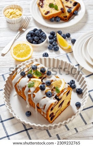 Lemon Blueberry Loaf with Lemon Glaze on dish on white wood table with ingredients on background, vertical view from above