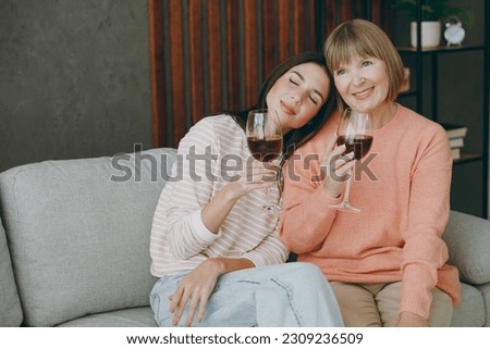 Two calm adult women mature mom young kid wear casual clothes drink wine put head on shoulder sit on gray sofa couch stay at home flat rest relax spend free spare time in living room. Family concept