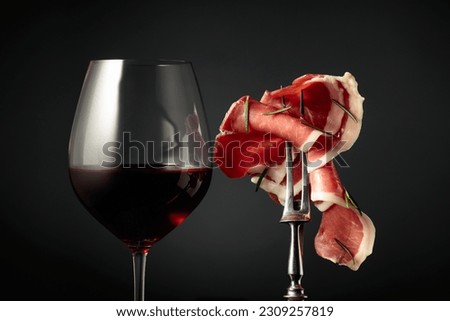 Glass of red wine and sliced prosciutto with rosemary on a fork, black background.
