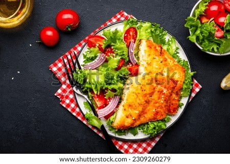 Fried white fish fillet with vegetable salad from lettuce, cherry tomatoes and red onion, black table background, top view