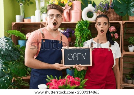 Young hispanic man a woman working at florist holding open sign afraid and shocked with surprise and amazed expression, fear and excited face. 