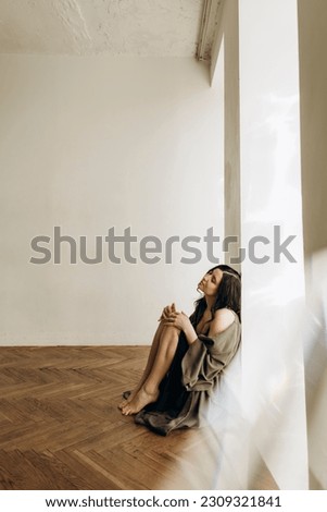 young beautiful woman sits on the floor with her back against the wall. a brunette girl sits on a parquet floor in a room with white walls and dreams about something.