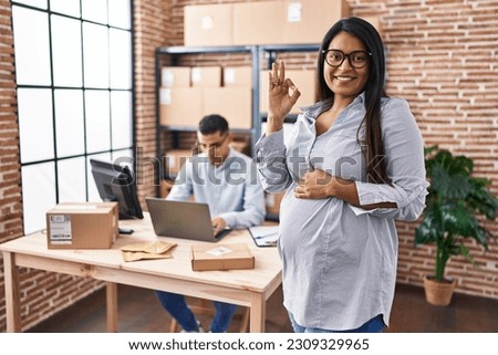 Young hispanic woman expecting a baby working at small business ecommerce doing ok sign with fingers, smiling friendly gesturing excellent symbol 