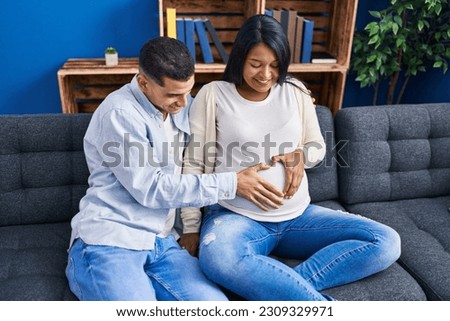 Young latin couple expecting baby touching belly doing heart gesture at home