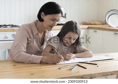 Loving engaged grandma and focused cute girl drawing cartoon sketches in paper album together, sitting at kitchen table, using colorful pencils, training kids creativity