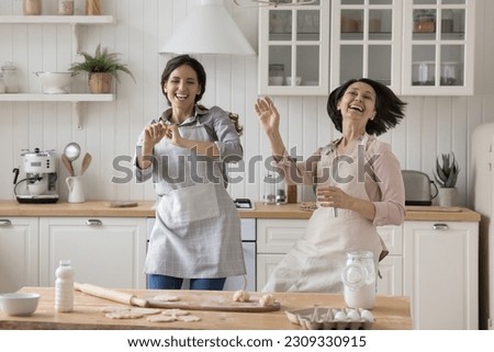 Cheerful adult daughter and excited mature mom in cooking aprons dancing at kitchen table with baking ingredients, laughing, having fun, enjoying music, movement, family culinary activity Royalty-Free Stock Photo #2309330915