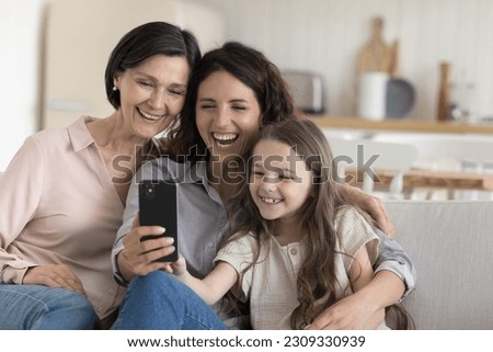 Joyful kid girl, mom, grandma relaxing on home sofa with smartphone, using app, media service for photo, taking selfie picture, enjoying domestic Internet communication