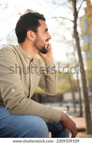 Side portrait of smiling young arabic guy talking on cell phone sitting outside
