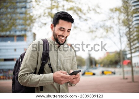 Portrait of handsome young middle eastern man walking outside with bag and using cell phone Royalty-Free Stock Photo #2309339123