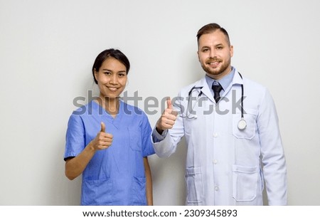 Young caucasian doctor and asian  physical therapist stand with finger thumb up in front of white wall.