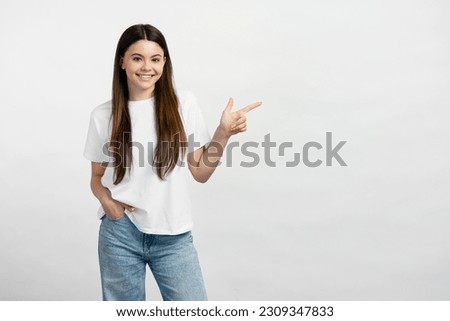 Smiling  teenage girl wearing white t shirt and stylish jeans, pointing finger at copy space, place for text. Happy teen showing to empty place, advertisement. Shopping concept 